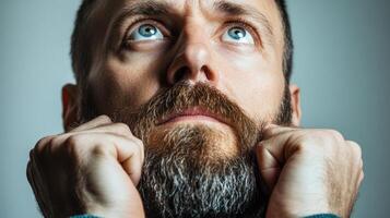 A man with a deep expression is pulling at his beard, his gaze upward, reflecting deep thought or concern photo