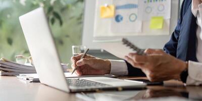 Elderly businessman reviewing data on a laptop while taking notes in a notebook at a modern office. photo