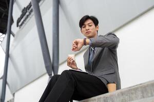 Young Asian businessman checking the time while enjoying a snack in a modern outdoor business setting. photo