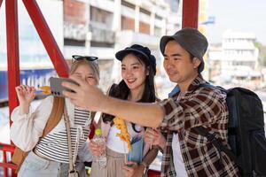 Young Asian friends taking a selfie while enjoying street food at a vibrant market, celebrating travel and friendship. photo
