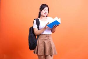 A person with a backpack and a book posing in front of a solid orange colored background photo