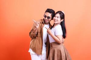a couple asia of teenage lovers cheerful expression pointing right where the man and woman stand facing each other romantically. isolated on orange background photo