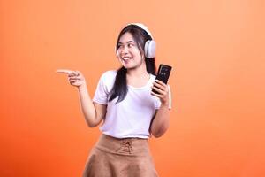 Smiling woman with headphones holding phone pointing to the left on orange backdrop photo