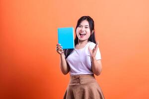 A person holds a blue book with a happy expression while on a solid orange background is visible photo