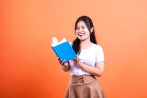 A woman holds a blue book and laughs while standing against a solid orange background in a studio setting photo