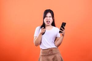 Furious woman pointing at the viewer while holding a cell phone isolated on an orange background photo