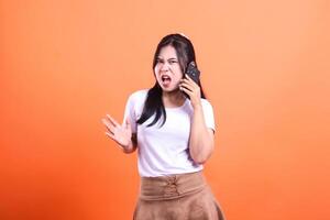 Young woman with long black hair in a white t-shirt talking on a cell phone with open mouth isolated on an orange background photo
