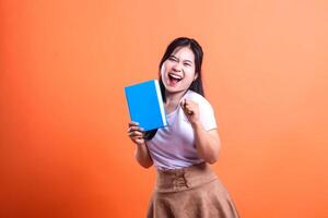 Excited woman holding a blue book against an orange backdrop with a victorious gesture ready to start reading it photo