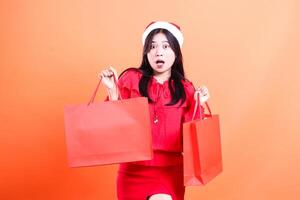 gesture of girl standing wearing red blouse and santa hat, standing very surprised to the front with both hands holding red bag of christmas presents to the front and left, isolated orange background photo