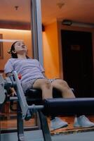 A woman uses a leg extension machine in a fitness center. She sits with her legs on the bar of the machine. The background is a window with lights, a door and some equipment. photo