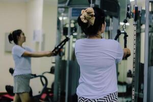 A woman asia with her back facing forward is using a cable machine, holding the handles, and exercising at a gym. The scene focuses on physical activity and fitness. photo