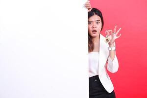 A woman is holding a house key and hiding behind a white banner with a surprised expression, isolated on a red background photo