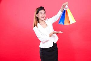 A woman holds three colorful paper shopping bags with her arm raised, while the other hand is showing an open palm, isolated on a red background photo