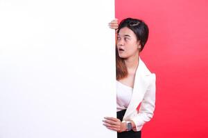 A woman wearing a white jacket and a black skirt is holding a large white cardboard while looking to the side, isolated on a red background photo