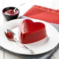 A red pudding shaped like Heart shape is placed on a white plate with a small spoon next to it, displayed on a clean background photo