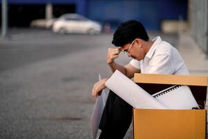 A man sits on the ground next to a cardboard box with papers in it. He looks upset and is rubbing his forehead photo