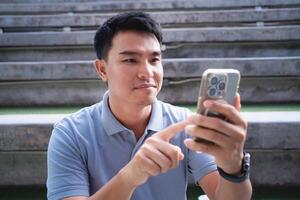 A man is sitting on a bench and looking at his phone. He is pointing at something on the screen photo