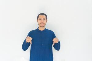 Muslim men wearing blue baju koko is pointing down and smiling in isolated white background photo