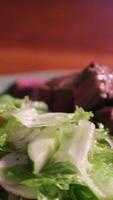 Fresh salad with green lettuce leaves and sliced tomatoes topped with grated cheese on white plate. Grilled piece of meat under vegetables served with floral design. video