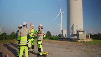 Four workers discuss plans while studying a blueprint at a wind farm. They wear safety gear and helmets, showcasing teamwork in a renewable energy setting. video