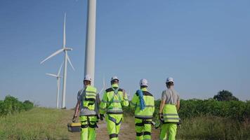 Group of four workers in safety gear review plans at a wind turbine installation location under clear skies. They are focused on their task while ensuring safety standards. video