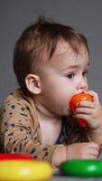 Cute Caucasian baby boy lies on the carpet in the room. Adorable child pulling his toys into his mouth. Blurred backdrop. Vertical video