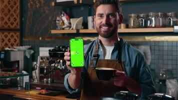Glad Caucasian man barista small business entrepreneur male demonstrating mobile phone green screen smartphone application in cafe employee guy worker hold coffee drink cup restaurant online app tech video