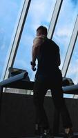 Sportsman on a treadmill indoors on sky background. Fitness guy is running on a simulator in the gym and looking through windows. Rear view. Vertical video