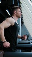 Sportsman is running on a treadmill. Young bodybuilder exercising on a simulator on the background of modern interior of fitness center. Profile view. Vertical video