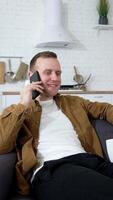 vrolijk jong Mens pratend de telefoon Aan bank. vent is houdende Aan bankstel met een kop van koffie en hebben een aangenaam gesprek door mobiel telefoon Bij huis. verticaal video