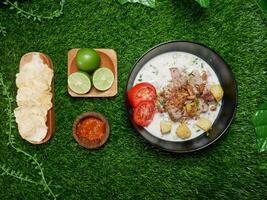 Top view Soto Betawi dish with coconut milk broth, beef, potatoes, and fried shallots. Served with fresh tomato slices, spicy sambal, crispy crackers, and fresh limes. With synthetic green grass base photo