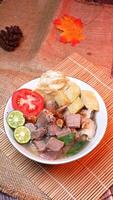 Close-up of a bowl of clear beef soup with chunks of beef, potatoes, fresh tomatoes, lime slices, and fried onions. Served on bamboo mats, burlap, and leaves. photo