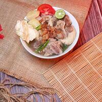 Top view of a bowl of oxtail soup or sop buntut with chunks of beef, potatoes, fresh tomatoes, lime slices, and fried onions. Served on bamboo mats, burlap, and leaves. Space for text available. photo