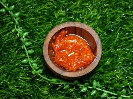 Indonesian traditional chili sauce named sambal terasi. crushed red chili peppers and mixed with shrimp paste, garlic, onion, and spices, served in a small wooden bowl on green synthetic grass base. photo
