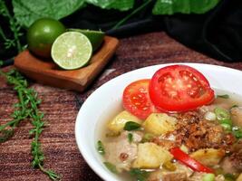 Selective focus clear beef soup served in a white bowl with a blurred background of lime, green leaves and black fabric, on a brown wood pattern background. photo