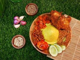 Top view A dish of cone-shaped yellow rice served with crispy fried chicken, fried vermicelli, spicy tempeh, potato sambal, fresh red sambal, cucumber slices, and spices like red onions and peppercorn photo