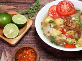 Close up of a bowl of beef soup with clear broth, potatoes, tomatoes and fried shallots, served with spicy red chili sauce and fresh lime. Served in a white bowl on a brown background photo
