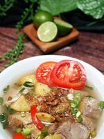 Clear beef soup served in a white bowl with a blurred background of lime, green leaves and black fabric, on a brown wood pattern background. Close up and Selective focus photo