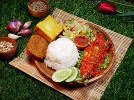 Close up A complete meal featuring white rice, fried fish with red sambal, fried tempeh, fried tofu, fresh vegetables, sambal, and cucumber slices. photo