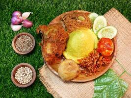 Delicious Yellow Rice with Traditional Indonesian Side Dishes, served with crispy fried chicken, fried vermicelli, spicy tempeh, red sambal, sambal egg, cucumber slices, potato fried on a straw base. photo