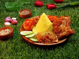 An authentic Indonesian dish of cone-shaped yellow rice, crispy fried chicken, fried vermicelli, sambal egg, cucumber slices, along with onions and spices on a wooden plate. photo