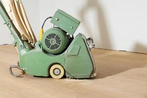 A retro green floor polishing machine with a fabric dust bag stands on a light wooden floor. Fine dust and a plain white wall are visible in the background. photo