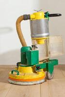 A green and yellow floor polishing machine with a cylindrical dust unit and hose sits on a worn wooden floor against a plain light colored wall. photo
