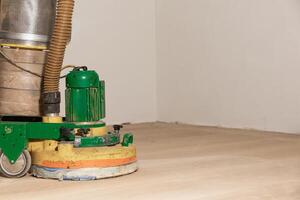 A green and yellow floor polishing machine with a dust collection system on a partially sanded wooden floor against plain off white walls. photo
