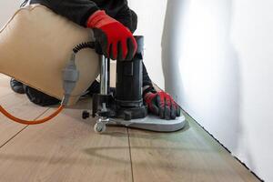 A worker kneels near a white wall, operating a floor polishing machine on light colored wooden flooring. An orange power cord connects to the machine. photo
