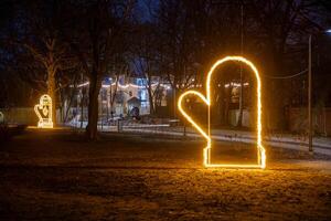 Illuminated mitten shaped lights line a pathway in a park at night, surrounded by bare trees. A well lit building with large windows is visible in the background. photo