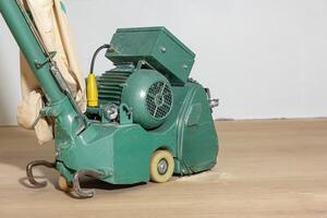 A green floor polishing machine with a motor, dust bag, and wiring on a light wooden floor. Partially polished surface and dust are visible. photo