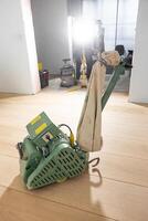 A green floor polishing machine with a beige dust bag on a light wooden floor. Construction tools and a work light are visible in the background. photo