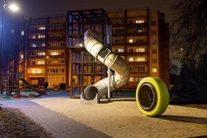 Nighttime view of a playground with a tall spiral slide, yellow play element, paved walkway, and a lit residential building in the background. photo