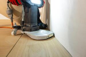 A person operates a floor polishing machine with a metallic base and bright light, smoothing a wooden floor near a white wall. An orange cord is visible. photo
